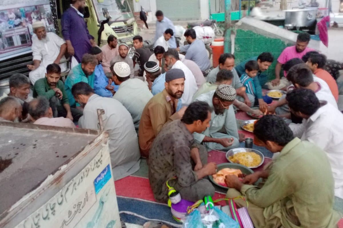 People seated on mats eating together, with cooking pots behind.