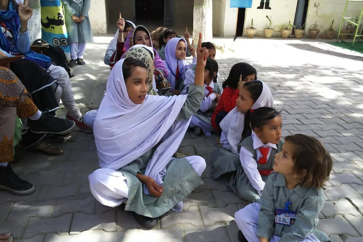 Girls seated outdoors during a class, one raising her hand to answer.