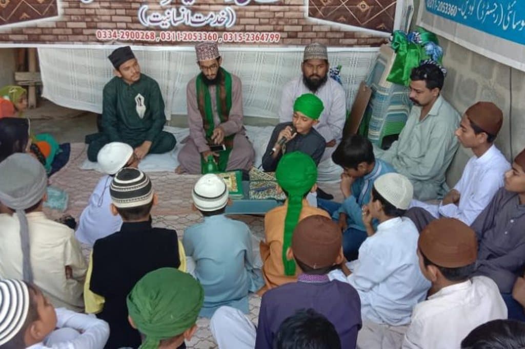A naat gathering with a boy reciting into a microphone, elders and children seated together.